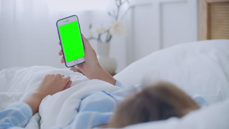 Young-woman-with-cell-phone-on-bed-shoulder-view.-Technology-addiction.-Green-screen-phone.-Young-woman-touching-belly-and-using-smartphone-in-bedroom